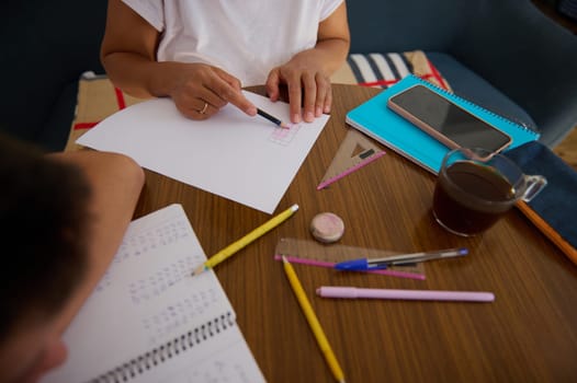 Two students work together at a home table, surrounded by notebooks, pens, and a smartphone. They engage in collaborative learning, drawing a diagram. The scene conveys focus and teamwork.