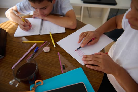 Two children working on school assignments at a wooden table, surrounded by various stationery items, illustrating concentration and focus in a home learning environment.