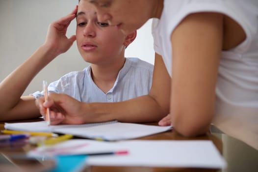 A mother assists her son with his homework at the kitchen table. The boy appears focused as he completes his assignment. Pencils and paper are scattered on the table around them.
