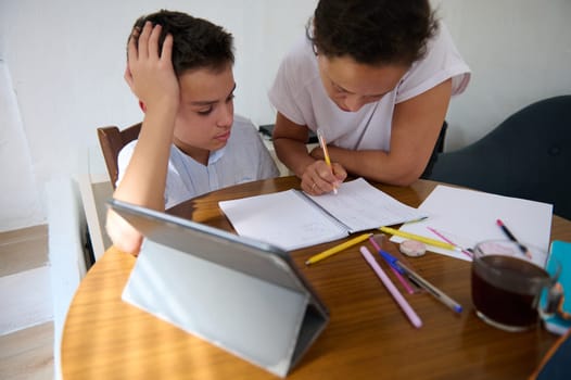 A mother assists her son with homework at a table, surrounded by study materials and a tablet. They focus on solving problems together.
