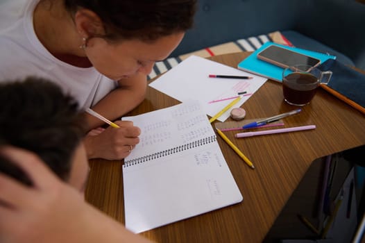 A mother assists her child with homework at a kitchen table, surrounded by stationery and a cup of coffee.