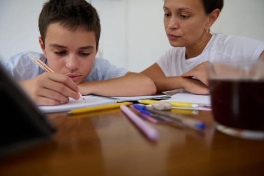 A mother assists her son with homework, guiding him at a wooden table filled with pens and paper. Their focus reflects a supportive learning environment, fostering education and understanding.