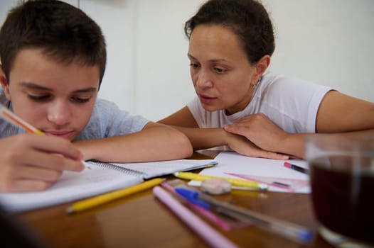A mother attentively helps her young son with his schoolwork at a kitchen table, surrounded by stationary. The setting portrays a nurturing and supportive home learning environment.