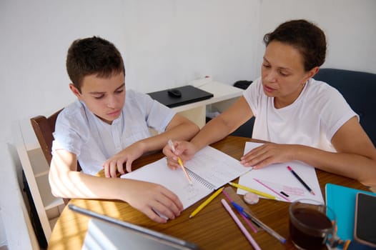 A mother assists her son with math homework at a table. They are engaged in solving problems, with notebooks and pencils scattered around. A laptop and a cup of tea are nearby.