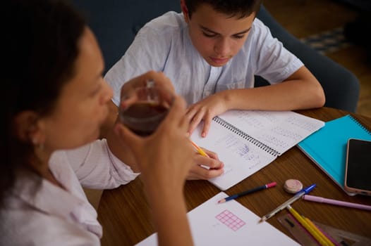A young boy concentrates on his math homework, jotting down notes in a spiral notebook. An adult, possibly a parent or tutor, sits nearby, providing support and guidance.