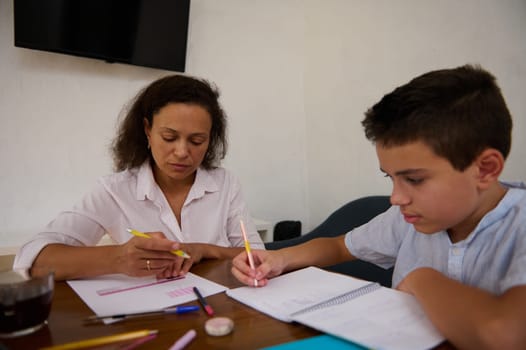 A mother and son sit together, deeply engaged in homework. They are indoors, surrounded by notebooks and pens, creating a warm learning environment.