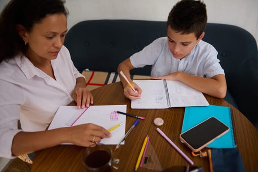 A mother and son sit together working on homework at a wooden table, surrounded by stationery. The mother guides her son with a ruler, depicting a supportive learning environment.