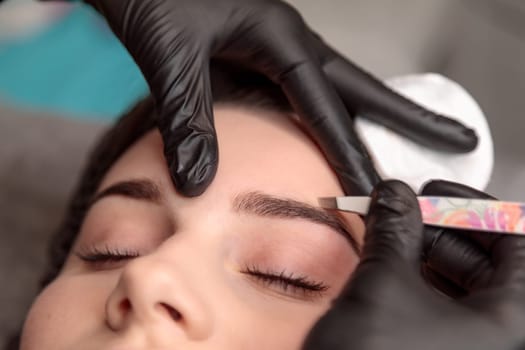 Beautician using tweezers to shape the eyebrows of a young woman in a beauty salon