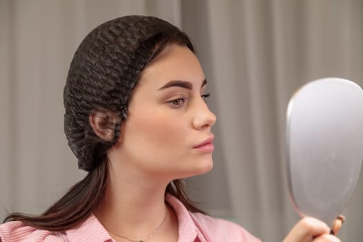 Young woman wearing hairnet looking at mirror checking eyebrows after microblading treatment in beauty salon