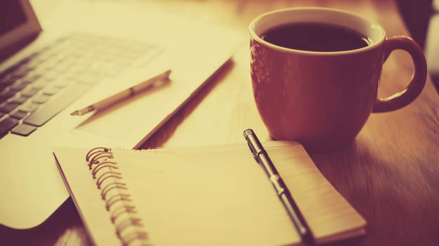 Cup of coffee sits on a desk with an open notebook and laptop computer, suggesting a break from work