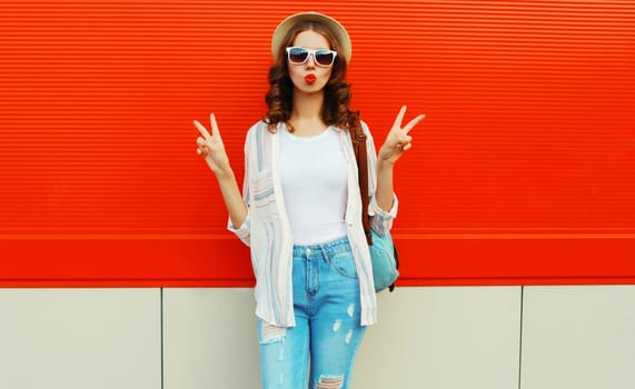 Portrait of beautiful young woman blowing kiss posing wearing a summer straw hat on red background