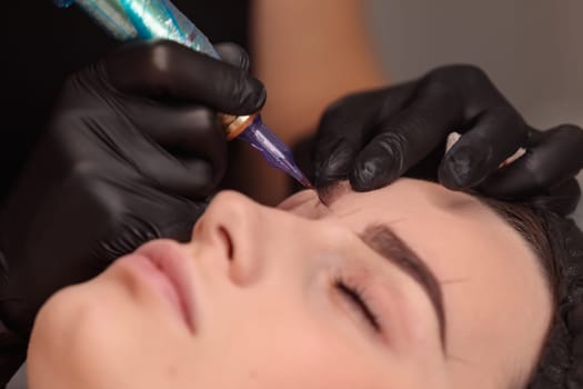 Cosmetologist making permanent eyebrow makeup using tattoo machine in beauty salon
