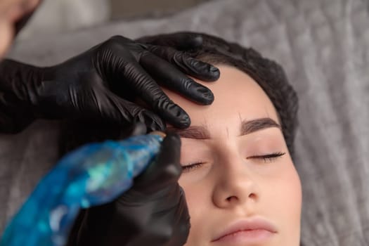 Beautician using tattoo machine applying permanent makeup on eyebrows of young woman in beauty salon