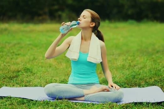 Fitness woman drinking water from bottle on a mat on the grass in summer park