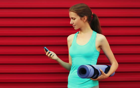 Fitness young woman with smartphone and yoga mat on red background