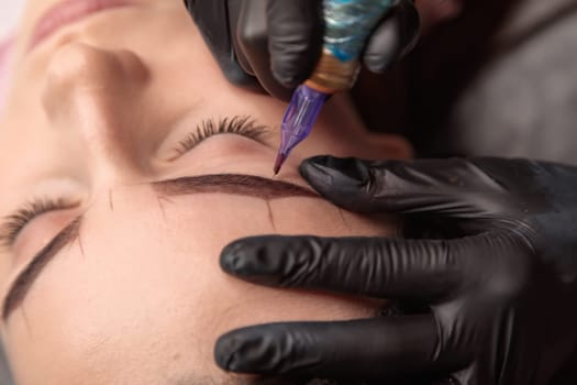 Beautician using tattoo machine for applying permanent makeup on eyebrows of young woman in beauty salon