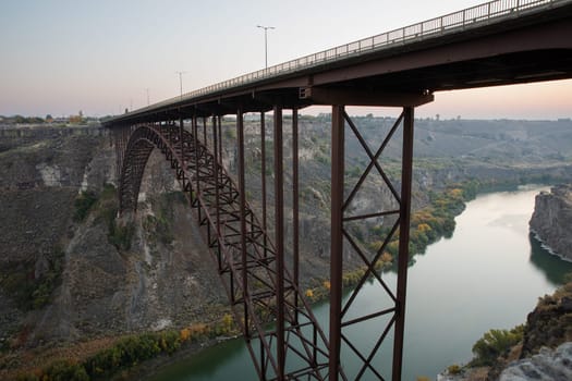 A magnificent steel bridge elegantly stretches across a tranquil river valley at dusk, framed by rugged cliffs and calm waters as seen from twin falls idaho
