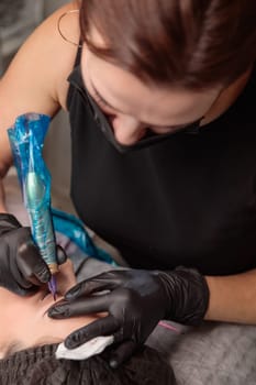 Beautician performing microblading procedure on customer's eyebrows using a tattoo device