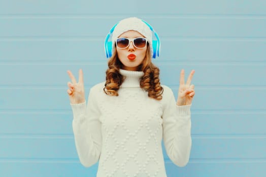 Fashion woman in headphones listens to music wear glasses, hat, sweater over blue background