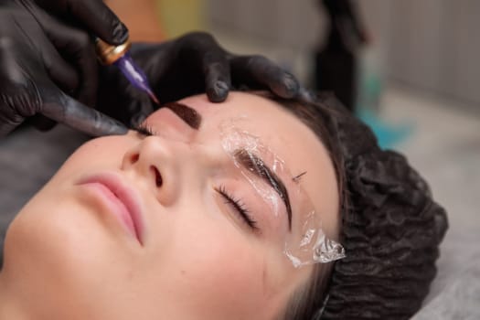 Beautician performing microblading procedure on eyebrows of young woman lying with closed eyes in beauty salon