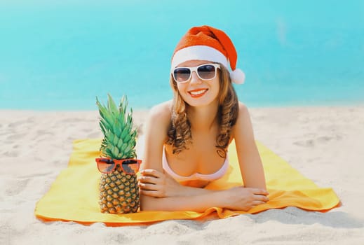 Christmas portrait young smiling woman in red santa hat and pineapple lying on beach on sea