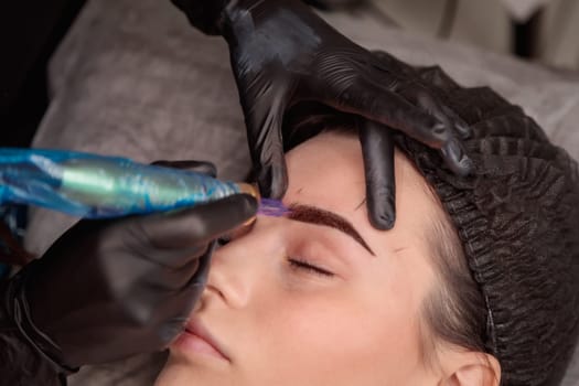 Cosmetologist making permanent eyebrow makeup using tattoo machine in beauty salon
