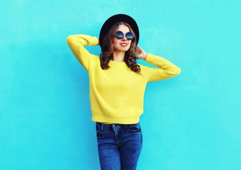 Fashion pretty woman wearing black hat and yellow knitted sweater over colorful blue background