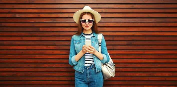 Portrait of young woman with phone in summer hat, backpack and jean jacket on wooden background