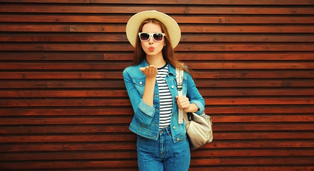 young woman blowing kiss with phone in summer hat, backpack and jean jacket on wooden background