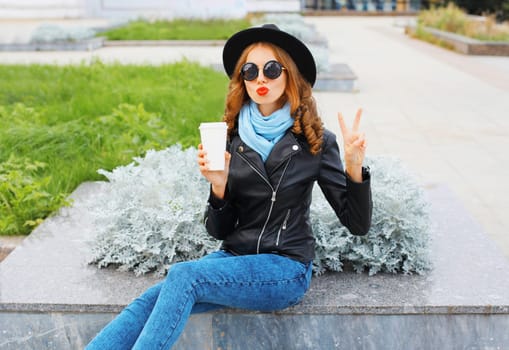 Fashion cool girl with coffee cup blowing a kiss wearing black rock jacket in city park
