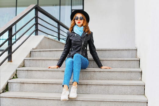 Fashion portrait pretty young woman wearing a black rock jacket and hat sitting in city