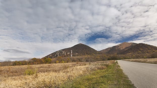 Serene landscape of mountains and open fields under a cloudy sky near a winding road in autumn season.