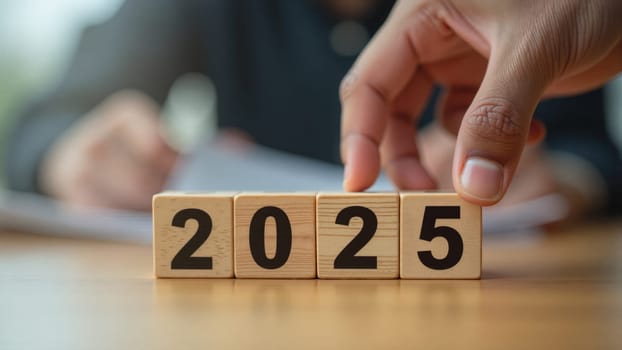 A hand carefully places a wooden block to highlight the year 2025, symbolizing aspirations and future plans. The backdrop reveals a focus on thoughtful preparation and new beginnings.