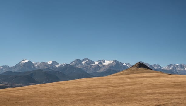 Breathtaking views of snow-capped peaks rise majestically in the background, while golden fields stretch across the landscape under a brilliant sky.