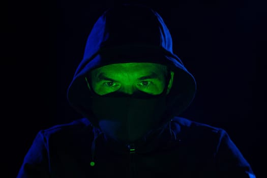 Short of computer hacker wearing white mask stealing data from a laptop. Masked hacker using laptop computer for organizing massive data breach attack on corporate servers in neon room.