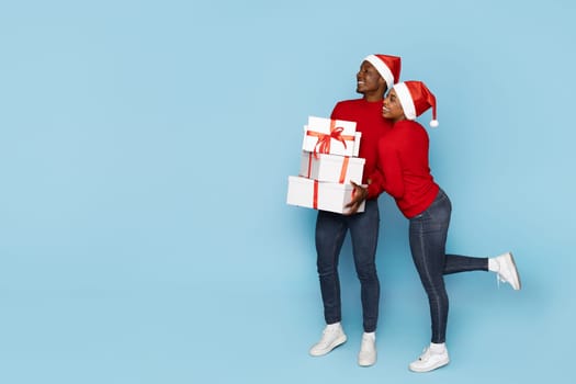 A cheerful couple in matching red sweaters and Santa hats embraces while holding beautifully wrapped gifts against a light blue background, embodying holiday spirit and joy, copy space