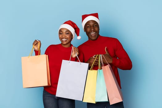A happy couple dressed in red sweaters and Santa hats holds colorful shopping bags. They are filled with excitement, embodying the holiday spirit as they celebrate Christmas.