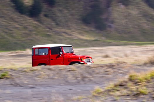 November 3, 2024. Bromo, East Java, Indonesia. A classic red Toyota Land Cruiser FJ40 drives through a scenic landscape with rolling hills and grasslands.