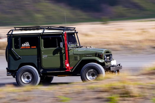 November 3, 2024. Bromo, East Java, Indonesia. A classic army green Toyota Land Cruiser FJ40 drives through a scenic landscape with rolling hills and grasslands.