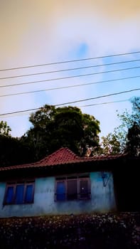 A rural house with a red-tiled roof surrounded by lush greenery under a serene, cloudy sky. The landscape conveys a sense of peace and rustic charm.