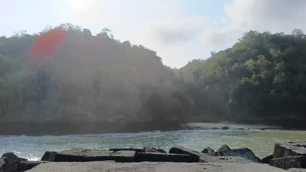 A rocky shoreline with a large rock formation in the background. The water is calm and the sky is blue