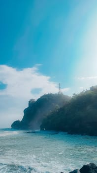 A rocky shoreline with a large rock formation in the background. The water is calm and the sky is blue