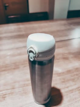 A silver thermos sits on a wooden table. The thermos is empty. The scene is simple and uncluttered, with the focus on the objects on the table