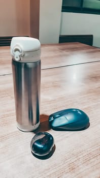 A silver thermos sits on a wooden table next to a computer mouse. The thermos is empty and the mouse is black. The scene is simple and uncluttered, with the focus on the objects on the table