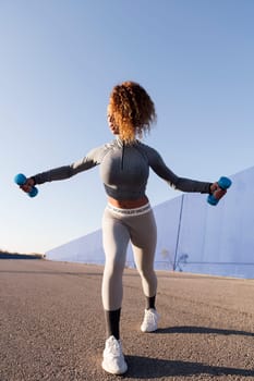 female athlete in sportswear working out with dumbbells during training on asphalt road, concept of urban sport and healthy lifestyle, copy space for text