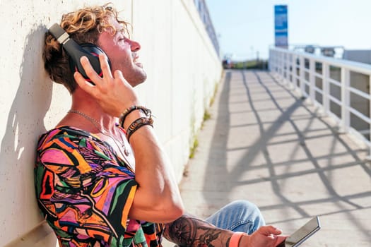 side view of dreamy male hipster sitting on street and enjoying songs in headphones with closed eyes