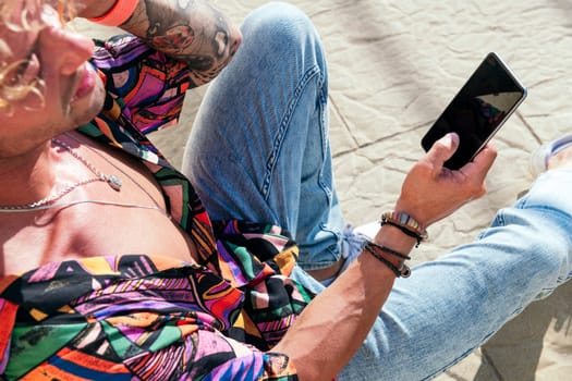 male cheerful hipster sitting in street and chatting on social media via mobile phone on sunny day