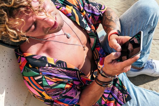 male cheerful hipster sitting in street and chatting on social media via mobile phone on sunny day