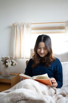 Smiling young woman reading a book by the Window, wrapped in soft blanket.