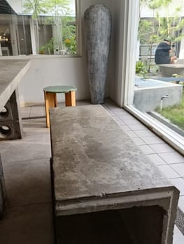A modern minimalist cafe interior featuring a large concrete chair and greenery. The bright space is filled with natural light, providing a serene and inviting atmosphere.